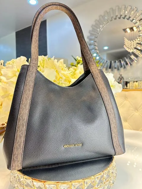 Bolso Michael Kors de cuero granulado negro, con asas y ribetes en cuero marrón con monograma, sobre una base de espejo junto a un ramo de flores blancas.